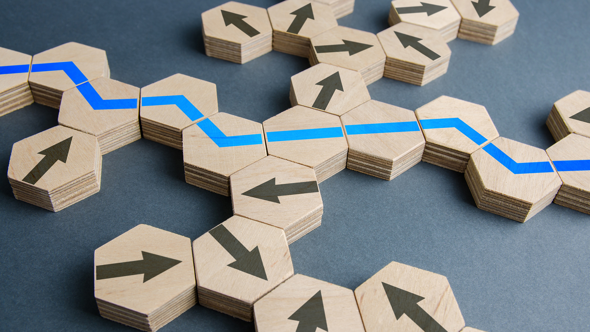 Wooden hexagonal tiles are arranged like a branching path on a dark blue surface, with some tiles showing black directional arrows and others forming a continuous bright blue line that zigzags across the route, suggesting navigation, choices, strategy, or decision-making.